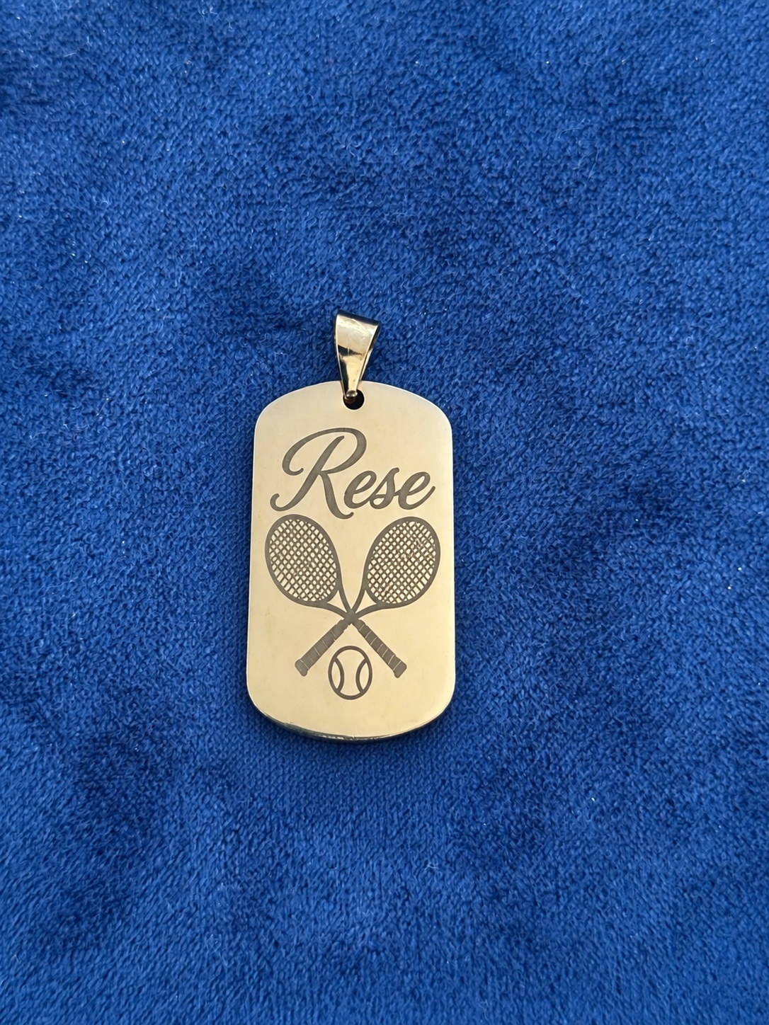 Gold-colored Rese dog tag engraved with crossed tennis rackets and a tennis ball on blue fabric